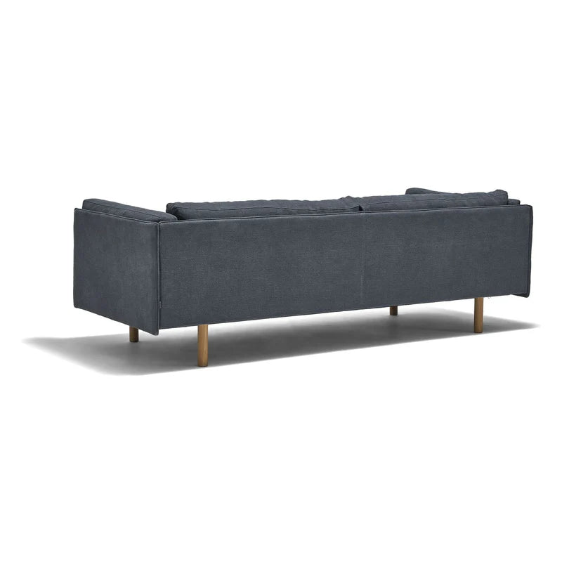A dark gray Sonder Sofa | Fabric by Wooden Stories, featuring a modern linear frame and wooden legs, is shown from the back at an angle. The sofa highlights clean Danish-inspired lines and three feather-filled back cushions, casting a shadow on the white background.