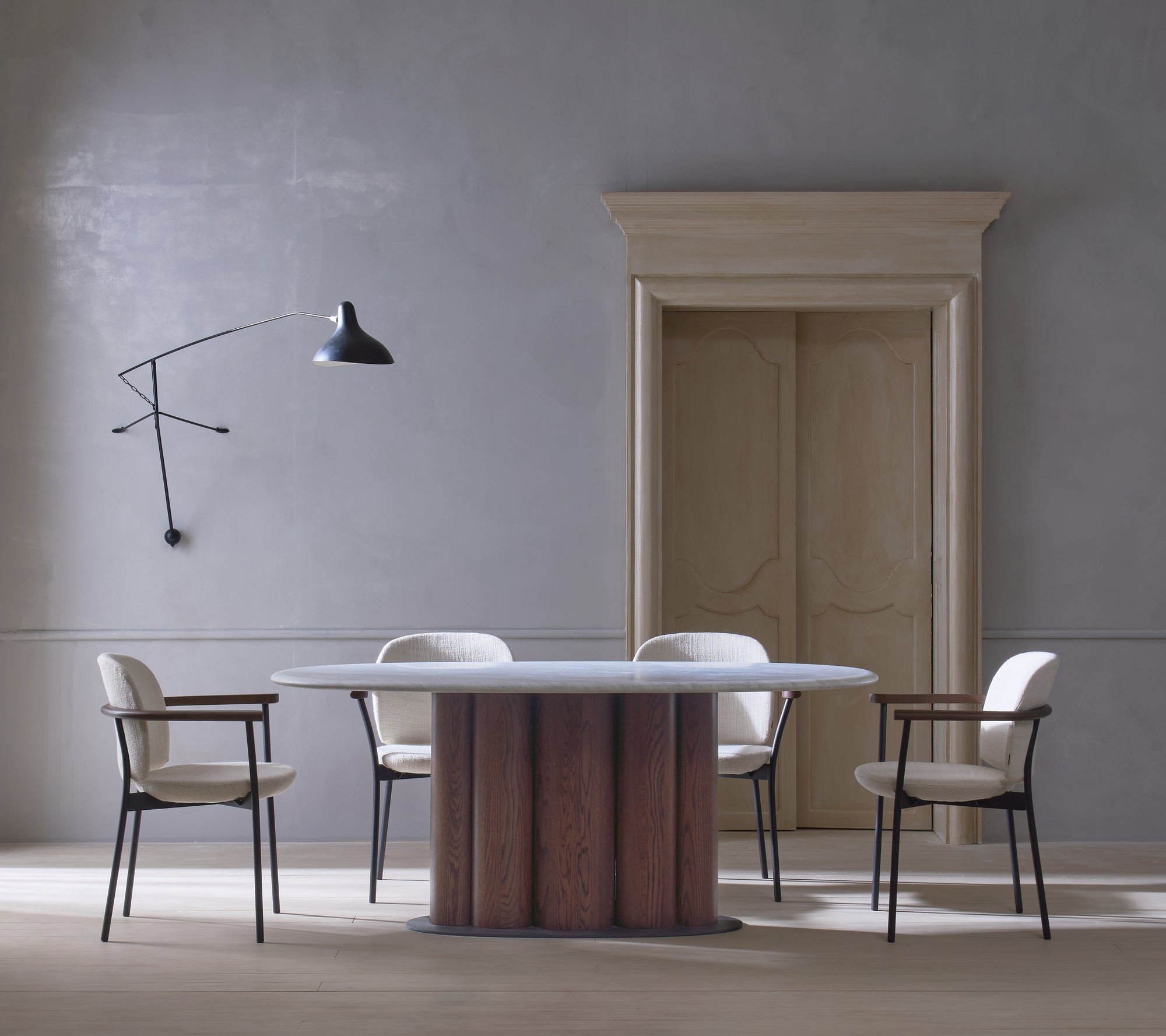 A modern dining area with a round marble-top table and wooden base, surrounded by four Wooden Stories Solace Dining Armchairs in fabric. A black wall-mounted lamp and tall wooden door complete the minimalist, neutral-toned room.