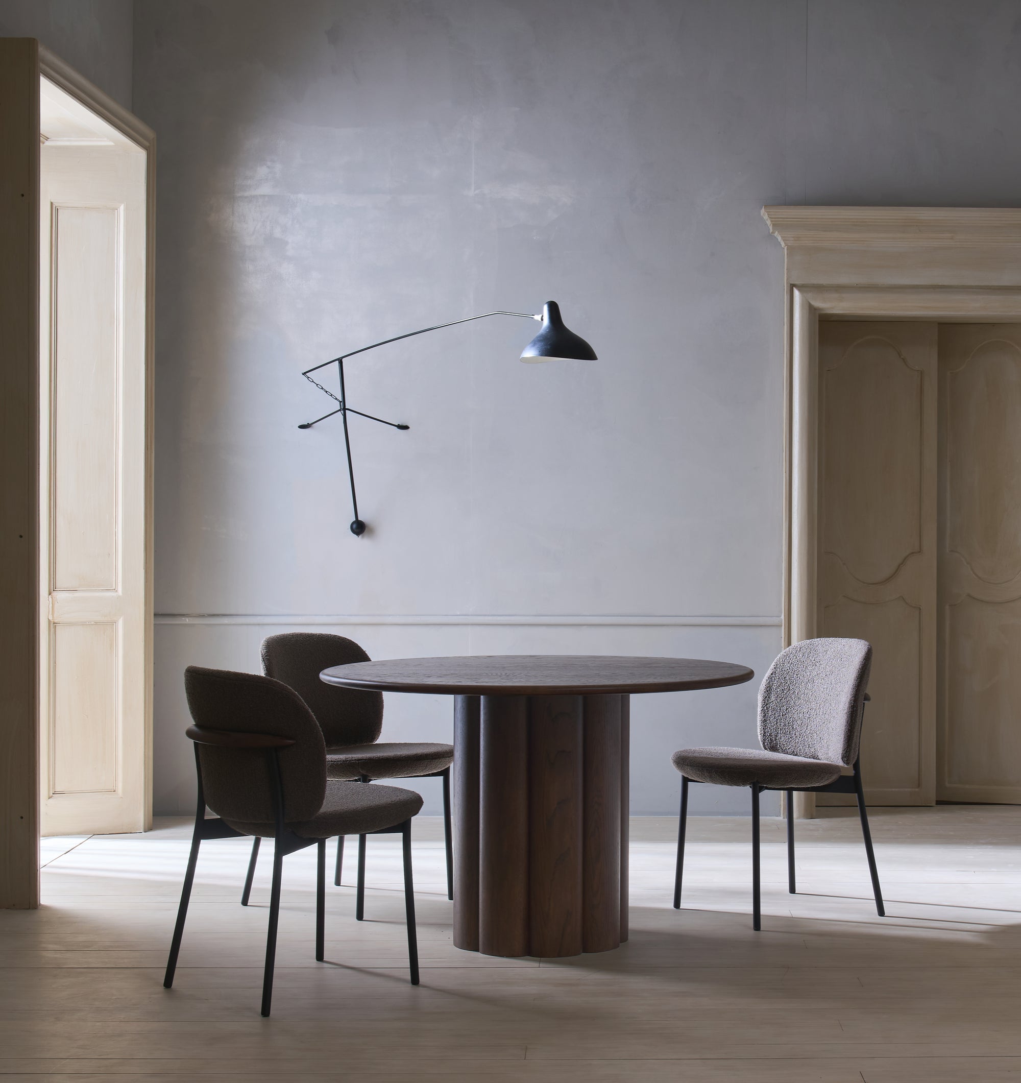 A minimalist dining room features a round wooden table and three Wooden Stories Solace Dining Chairs | Fabric, each with a premium steel frame, plus a modern black wall lamp. Light gray walls and tall beige doors complete the softly lit space.
