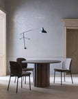 A minimalist dining room features a round wooden table and three Wooden Stories Solace Dining Chairs | Fabric, each with a premium steel frame, plus a modern black wall lamp. Light gray walls and tall beige doors complete the softly lit space.