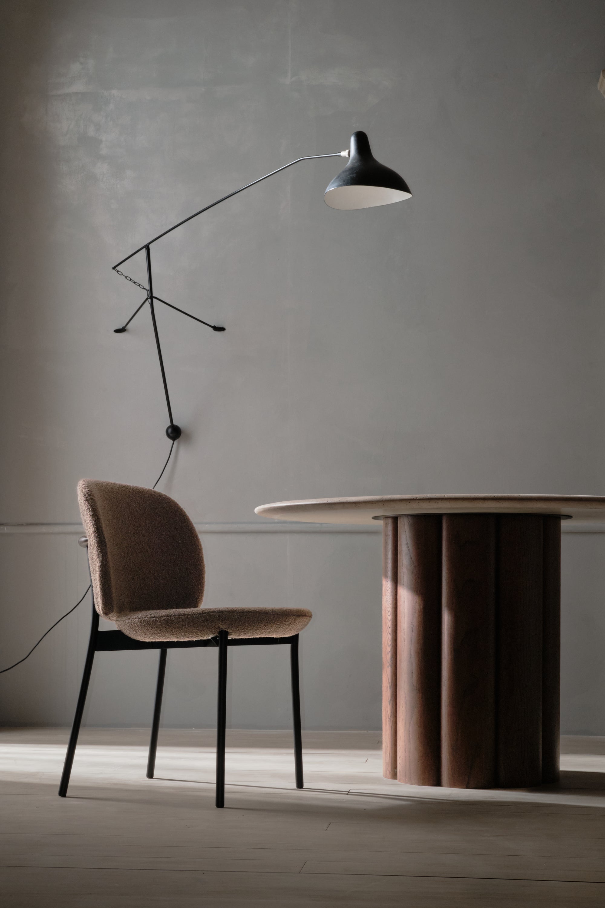 A minimalist room with a Wooden Stories Solace Dining Chair | Fabric, a round wooden table, and a modern black wall lamp against a plain gray wall. Natural light casts soft shadows on the floor.