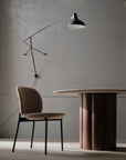 A minimalist room with a Wooden Stories Solace Dining Chair | Fabric, a round wooden table, and a modern black wall lamp against a plain gray wall. Natural light casts soft shadows on the floor.
