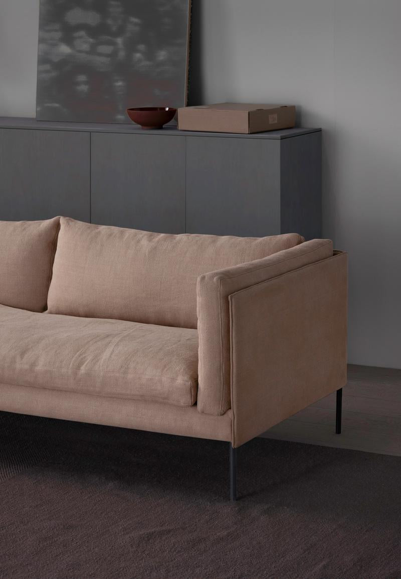 A modern beige Sonder Sofa by Wooden Stories, featuring thin black legs, is placed in front of a dark cabinet that displays a brown bowl, a rectangular box, and a framed abstract artwork against a light gray wall.