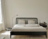 Minimalist bedroom with the Spindle Bed - Black Oak by Ethnicraft, beige bedding, two pillows, a black nightstand holding a lamp and books, light wood floors, and sunlight filtering through white curtains.