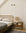 A minimalist bedroom featuring the Ethnicraft Spindle Bed in teak, white bedding, a gray blanket, a wooden nightstand with a lamp, books, decor accents, and two wall-mounted lights above the bed on a white wall.