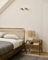 A minimalist bedroom featuring the Ethnicraft Spindle Bed in teak, white bedding, a gray blanket, a wooden nightstand with a lamp, books, decor accents, and two wall-mounted lights above the bed on a white wall.
