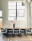 A minimalist dining room with a long solid ash table, six SOBU Sven Dining Chair - Black Ash, and a black vase with red branches, all beneath a large grid window filling the space with natural light.