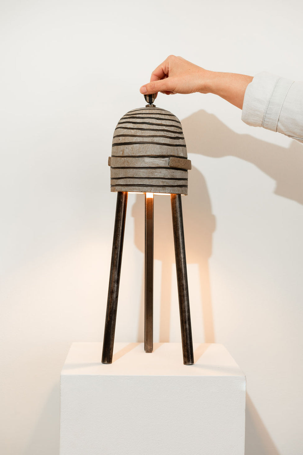 A hand lifts the lid of a modern, tripod table lamp made of textured, striped metal, standing on a white pedestal against a plain wall.