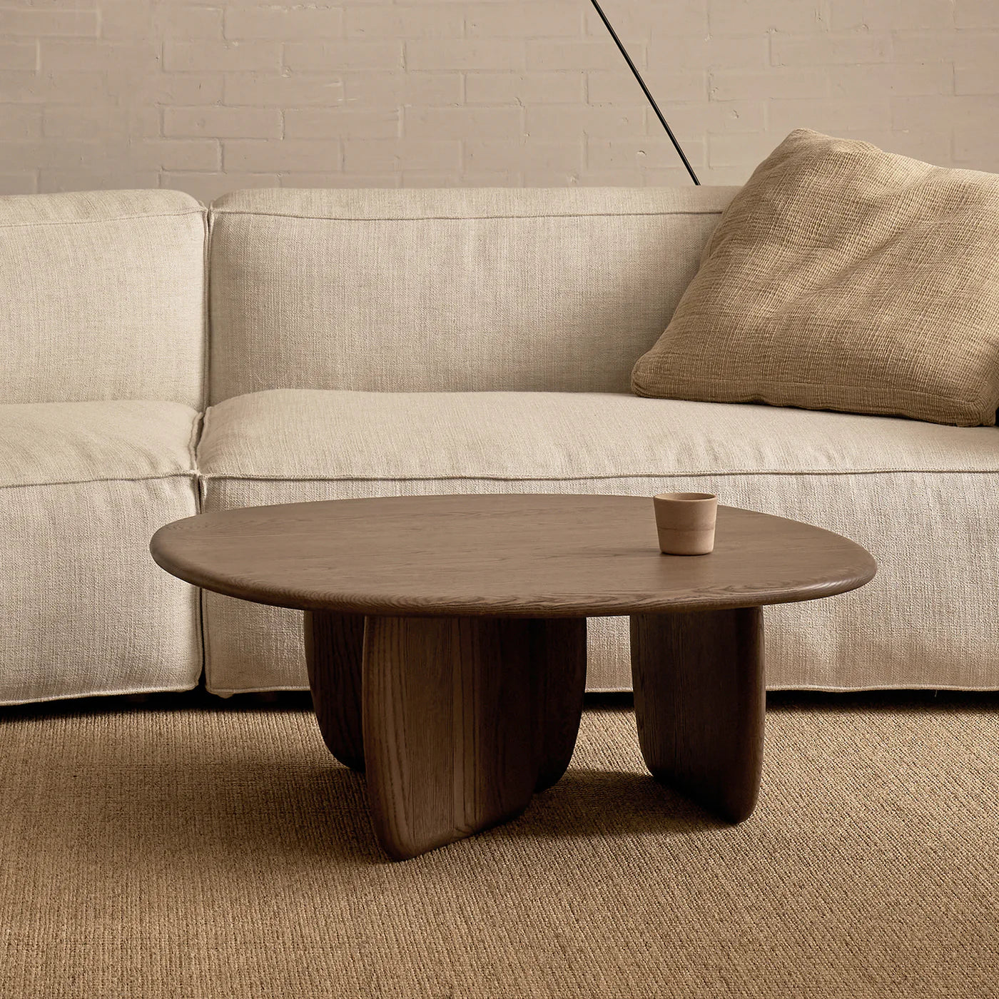 A modern living room with a beige sofa, textured beige pillow, the Wooden Stories Eden Coffee Table in Espresso Oak featuring sculptural timber legs, a small cup on the table, and a tan woven rug for an organic touch.