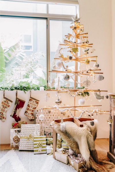 The SOBU Immortal Christmas Tree, a minimalist and sustainable wooden design, is decorated with ornaments and string lights, standing by the window among wrapped gifts, stockings, and cozy fur throws in a bright festive room.