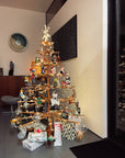 The SOBU Immortal Christmas Tree, a modern and minimalist wooden design, stands in the living room adorned with lights and ornaments. Wrapped gifts below glow in festive reflections from the nearby glass door.